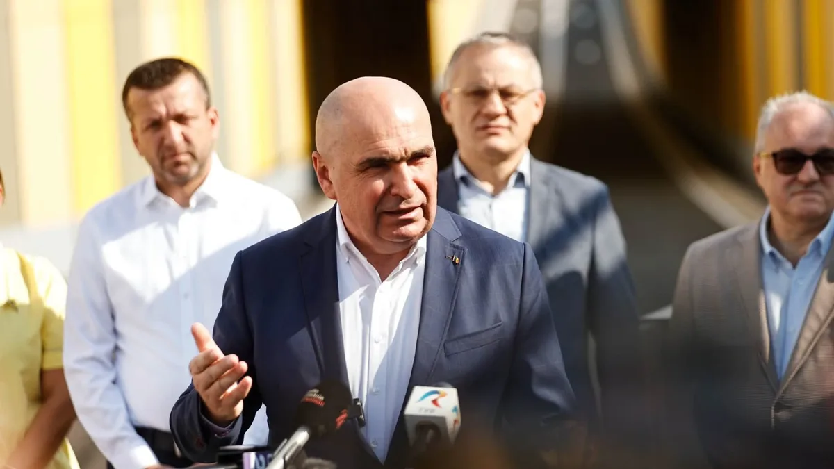 Ilie Bolojan gesticulează în fața microfoanelor, în aer liber, cu mai multe persoane în fundal.