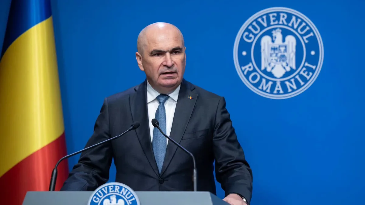 Ilie Bolojan la un pupitru cu două microfoane, lângă drapelul României, cu însemnele Guvernului în fundal.