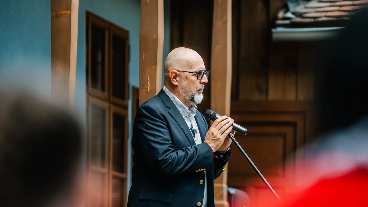 Kelemen Hunor vorbește la microfon, lângă stâlpi și uși din lemn, cu participanți neclari în prim-plan.