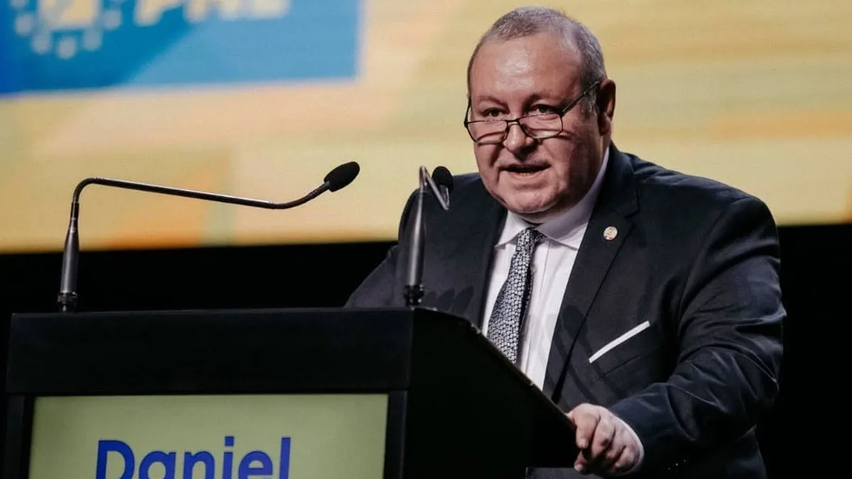 Daniel Fenechiu vorbește de la un pupitru cu două microfoane, pe un fundal galben și albastru.