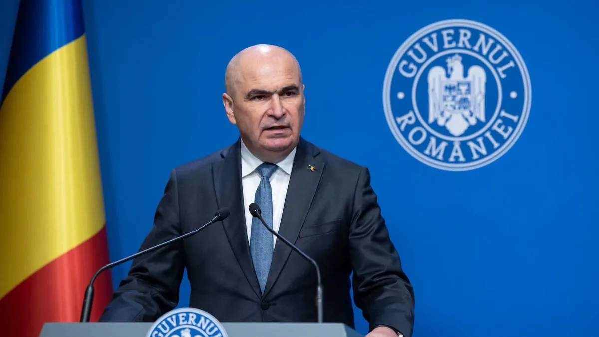 Ilie Bolojan la un pupitru cu două microfoane, cu însemnele Guvernului României și drapelul național în fundal.