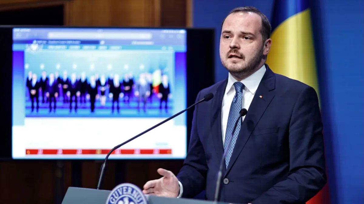 Alexandru Rogobete vorbește la un pupitru cu două microfoane, cu drapelul României și un ecran în fundal.