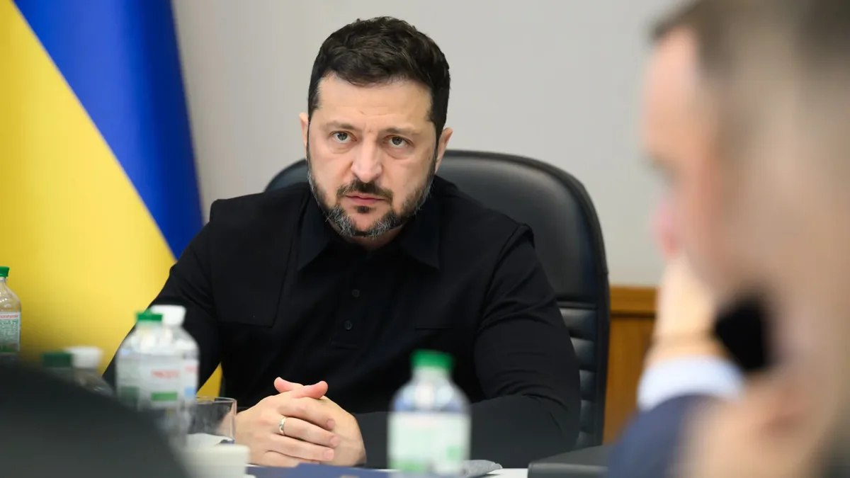 Volodîmîr Zelenski la o masă de reuniune, cu mâinile împreunate și drapelul Ucrainei în fundal.