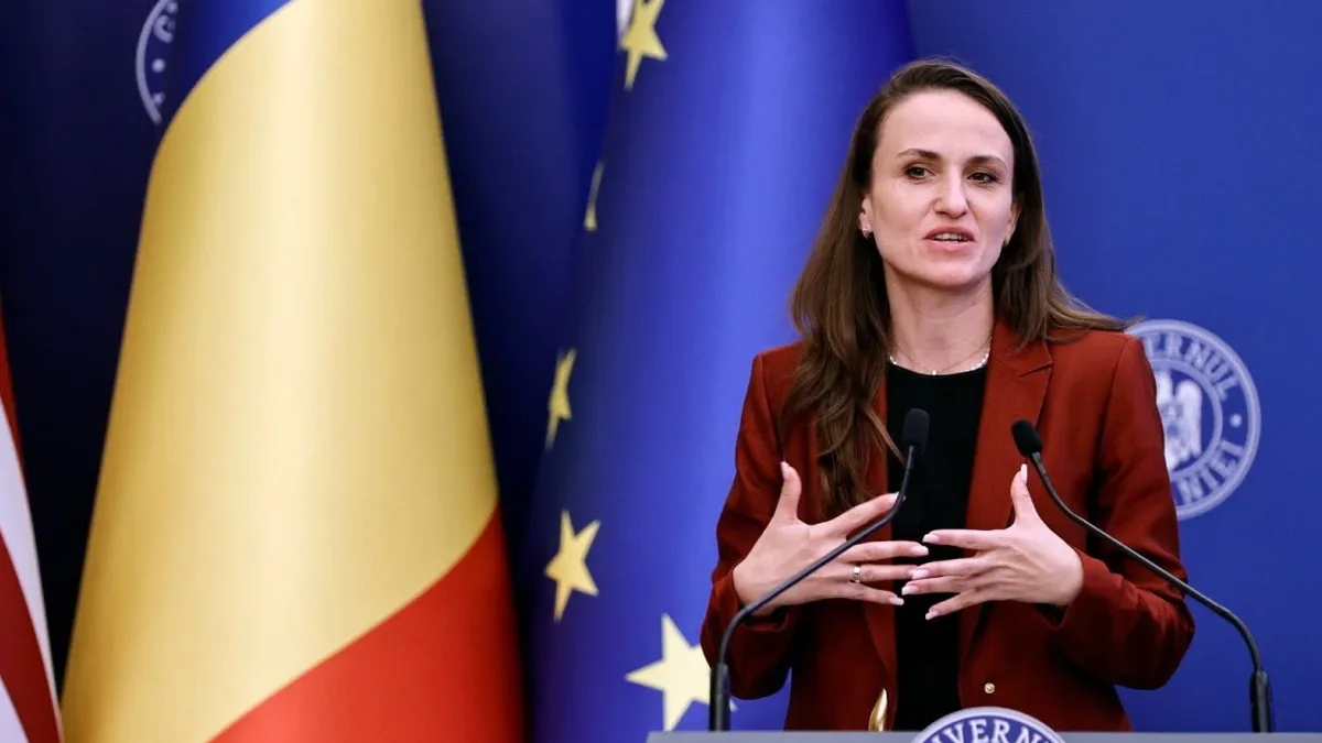 Oana Țoiu vorbește la două microfoane, în fața drapelelor României, Uniunii Europene și Statelor Unite.