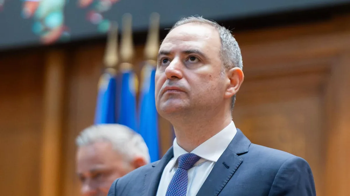 Bărbat în costum bleumarin și cravată albastră, în picioare într-o sală cu drapele în fundal.