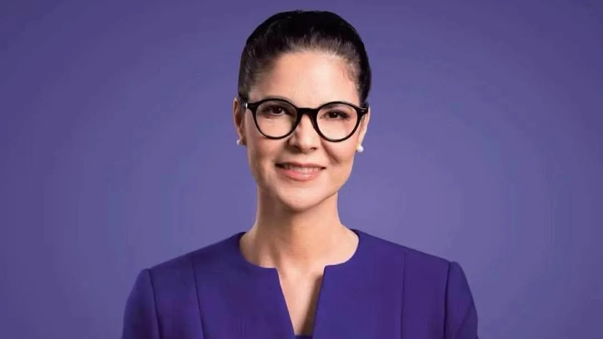 Ana Birchall, cu ochelari și sacou violet, fotografiată pe un fundal violet.