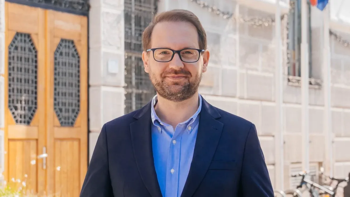Dominic Fritz, cu ochelari și sacou bleumarin, în fața unei clădiri cu fațadă de piatră și uși din lemn.