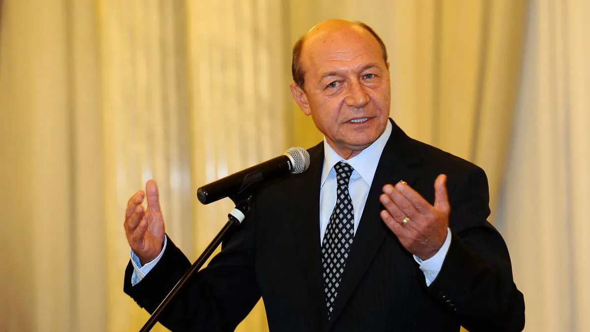 Traian Băsescu vorbește la microfon și gesticulează cu ambele mâini, în fața unor draperii.