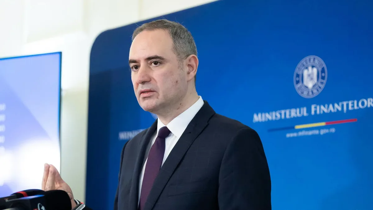 Alexandru Nazare vorbește la microfoane în fața unui panou al Ministerului Finanțelor.