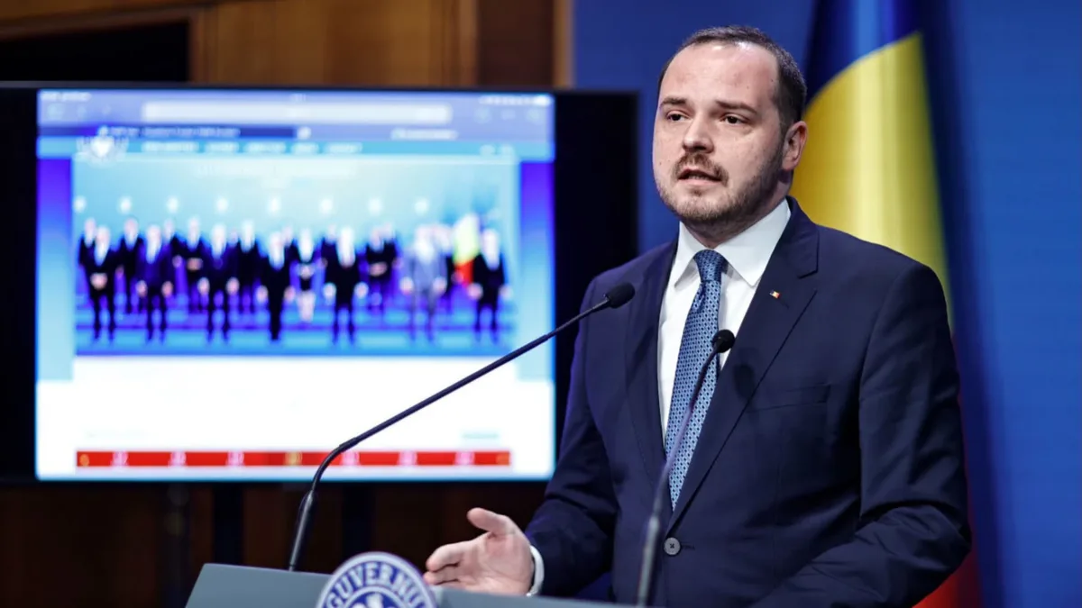 Alexandru Rogobete vorbește la un pupitru cu două microfoane, cu drapelul României și un ecran în fundal.