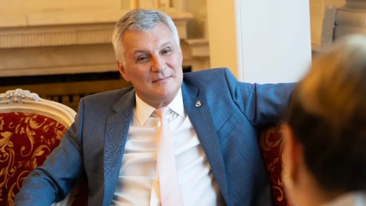 Daniel Zamfir, în costum albastru, așezat pe o canapea cu tapițerie roșie, în fața unei persoane văzute din spate.