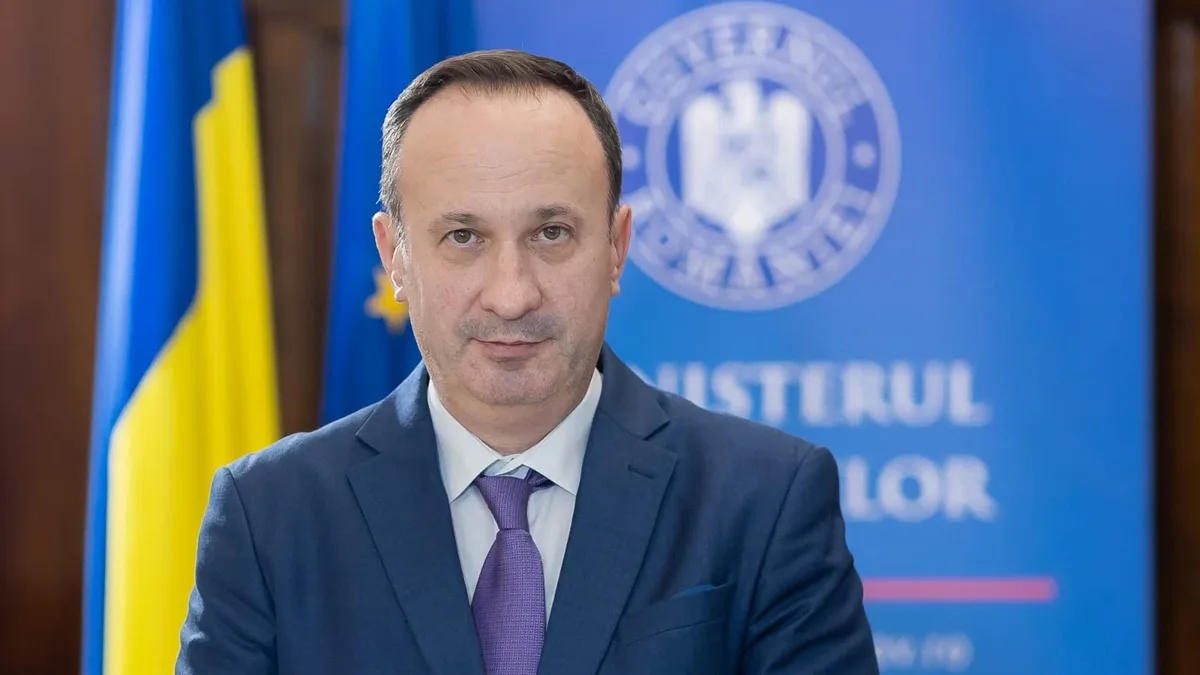 Adrian Câciu în costum albastru și cravată violet, cu drapele și un panou instituțional în fundal.