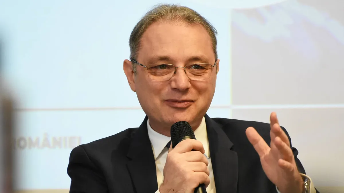 Luca Niculescu vorbește la un microfon ținut în mână și gesticulează în fața unui fundal luminos.