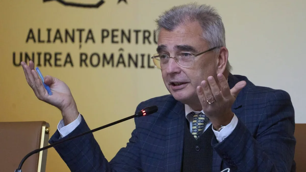 Un bărbat vorbește la microfon și gesticulează în fața unui panou cu numele Alianței pentru Unirea Românilor.