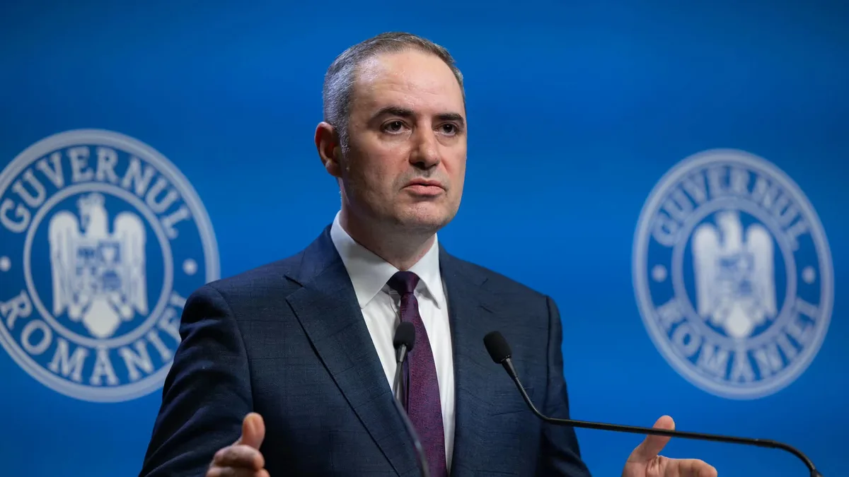 Alexandru Nazare gesticulează în fața a două microfoane, pe un fundal albastru cu însemnele Guvernului României.