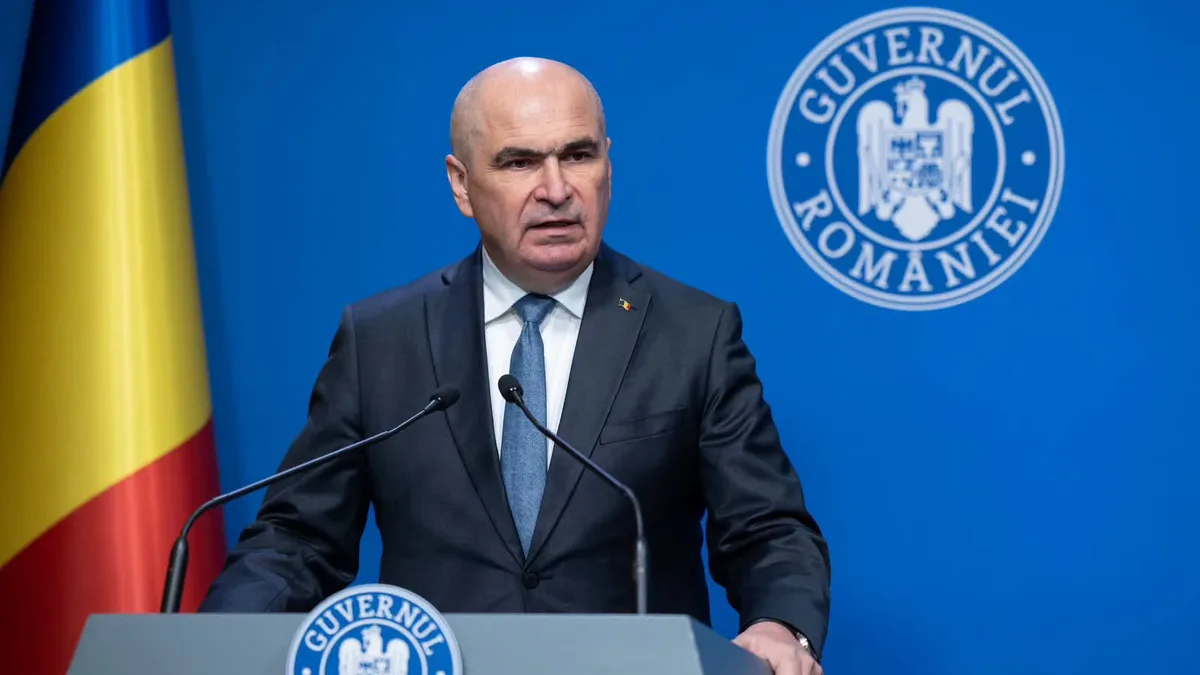 Ilie Bolojan la un pupitru cu două microfoane, lângă drapelul României și însemnele Guvernului.