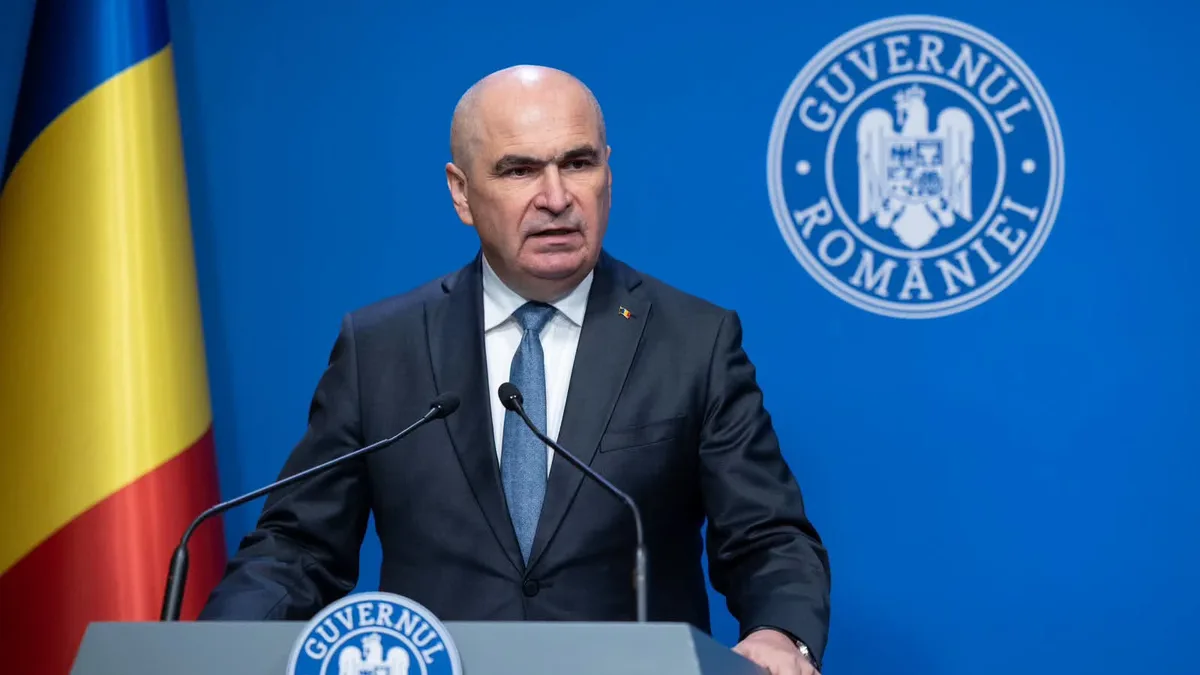 Ilie Bolojan la un pupitru cu două microfoane, lângă drapelul României, pe fundalul însemnelor Guvernului.