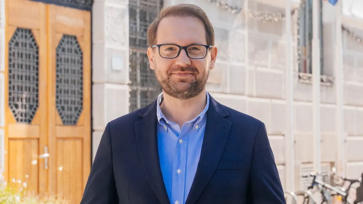 Dominic Fritz, cu ochelari, cămașă albastră și sacou bleumarin, în fața unei clădiri cu uși din lemn.