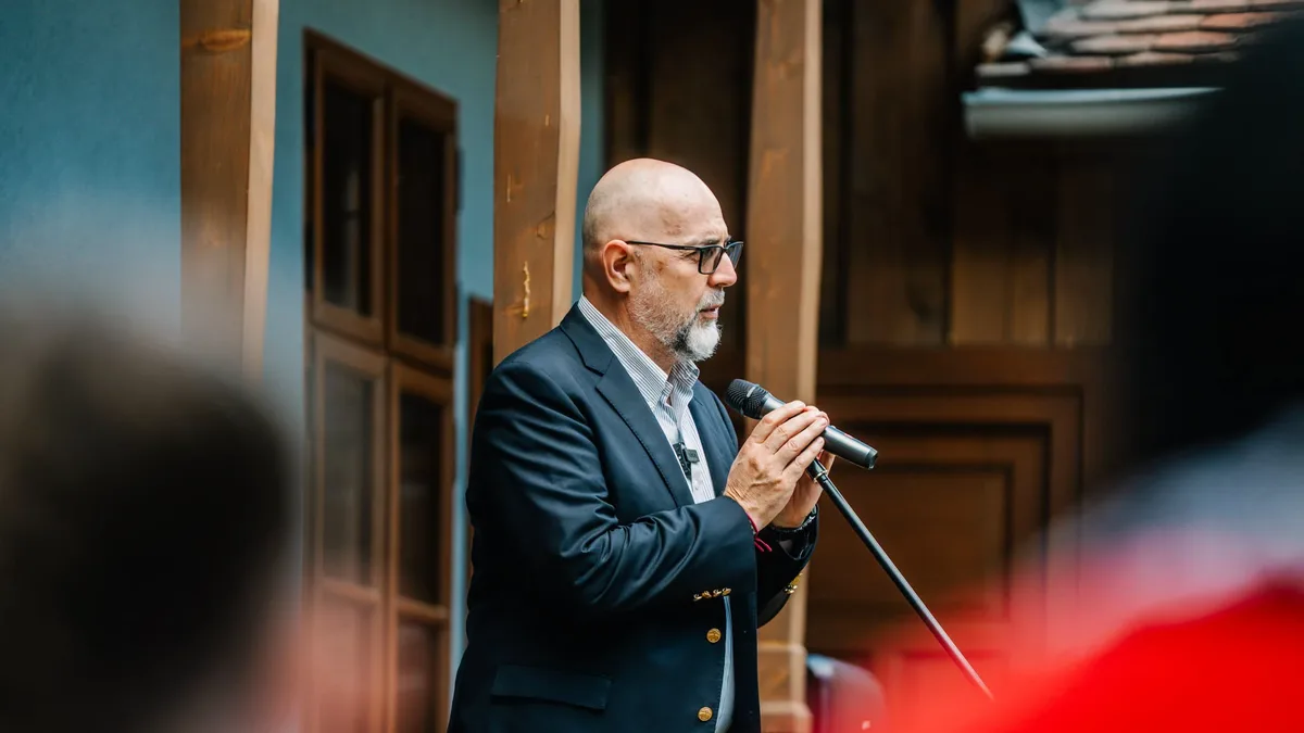 Kelemen Hunor vorbește la microfon, în picioare, lângă stâlpi și uși din lemn.