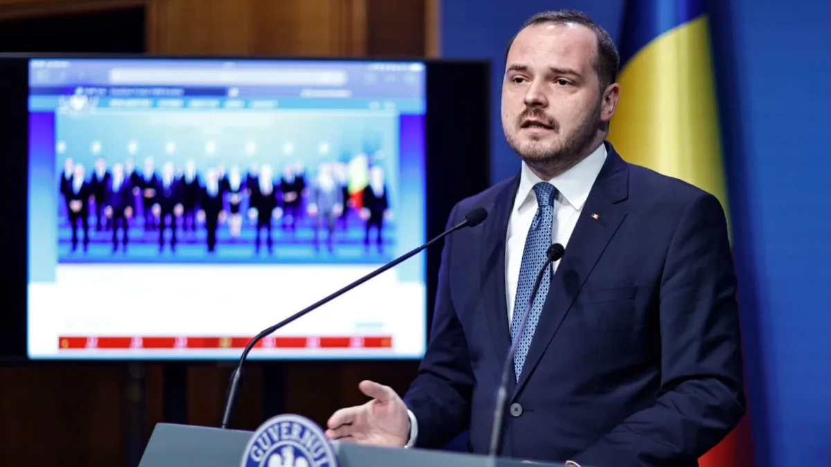 Alexandru Rogobete vorbește la un pupitru cu două microfoane, cu drapelul României și un ecran în fundal.