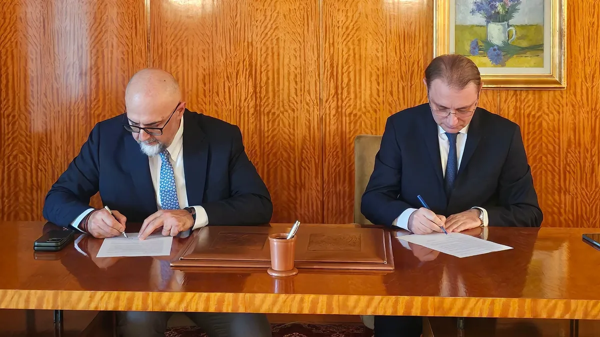 Doi bărbați în costume, așezați alături la o masă, scriu pe documente.
