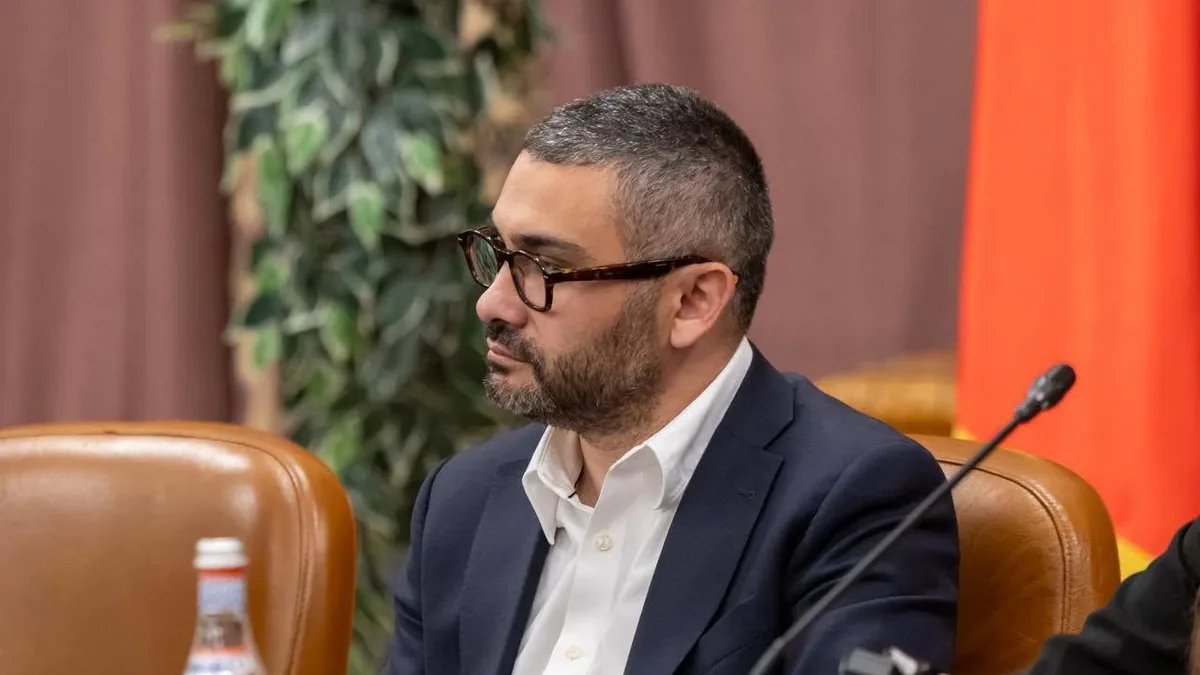 Florin Manole, în costum bleumarin și cămașă albă, așezat la o masă de ședință, lângă un microfon.