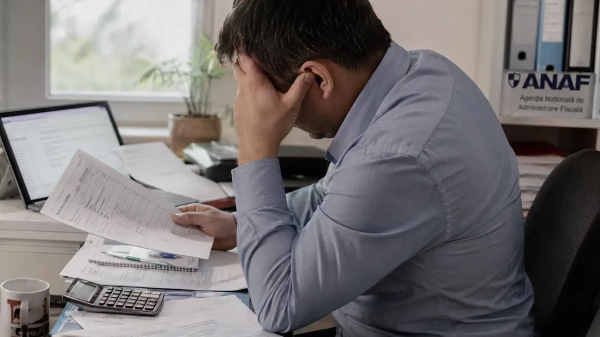 Un bărbat cu fruntea sprijinită în palmă ține un document la un birou cu laptop, calculator și hârtii; în fundal apare sigla ANAF.