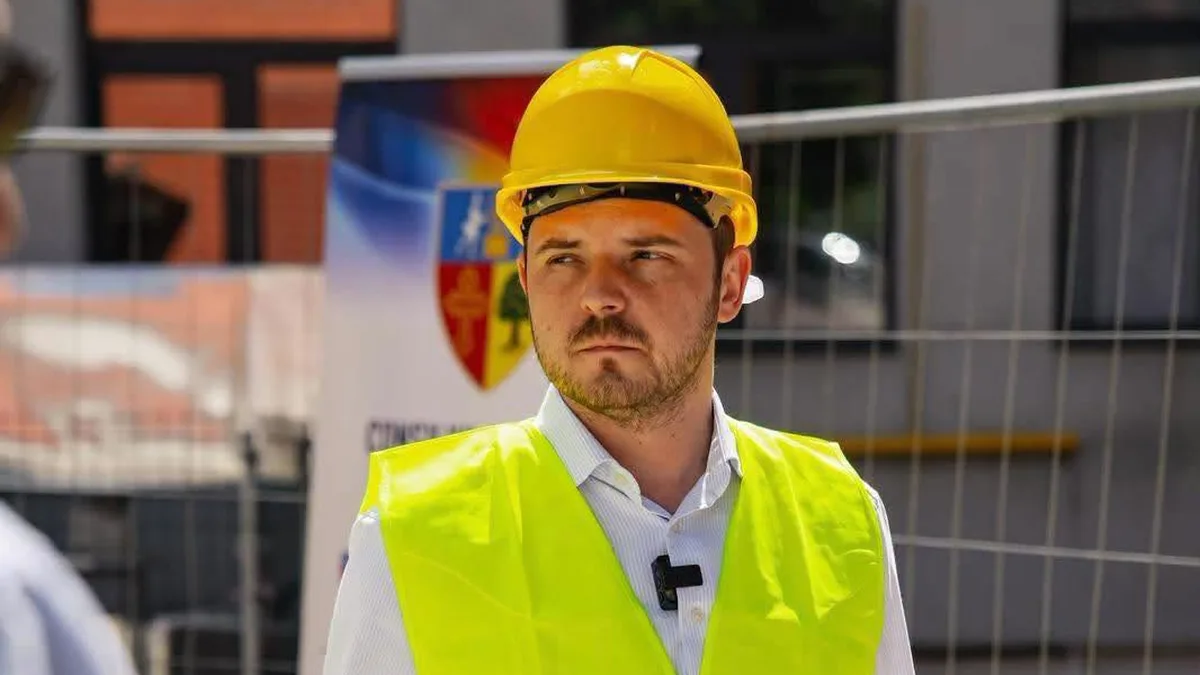 Alexandru Rogobete poartă cască galbenă și vestă fluorescentă, în fața unui gard metalic.