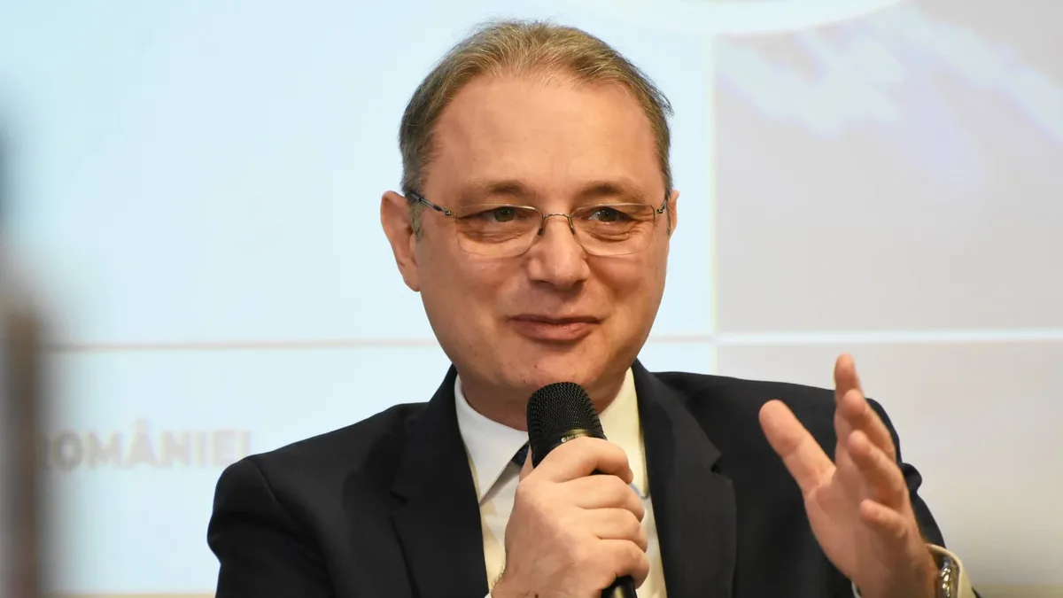 Luca Niculescu vorbește la un microfon ținut în mână și gesticulează în fața unui fundal luminos.