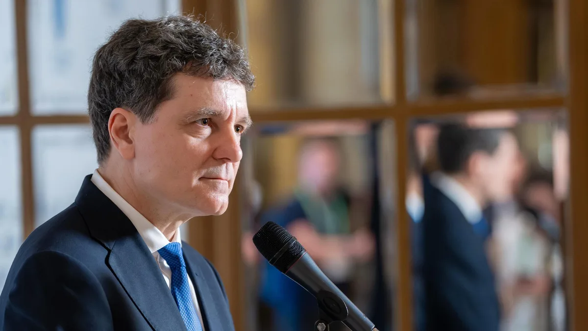 Nicușor Dan în fața unui microfon, într-un interior cu panouri vitrate și persoane neclare în fundal.