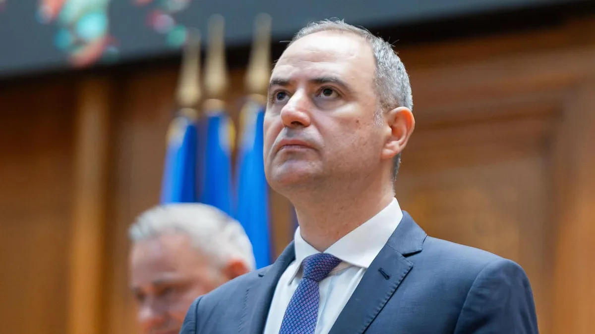 Alexandru Nazare, în costum bleumarin și cravată albastră, cu drapele neclare în fundal.