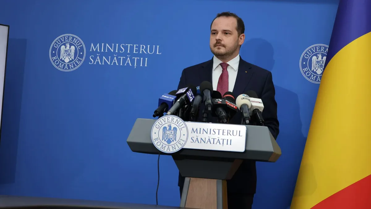 Alexandru Rogobete la pupitrul Ministerului Sănătății, în fața mai multor microfoane, cu drapelul României alături.