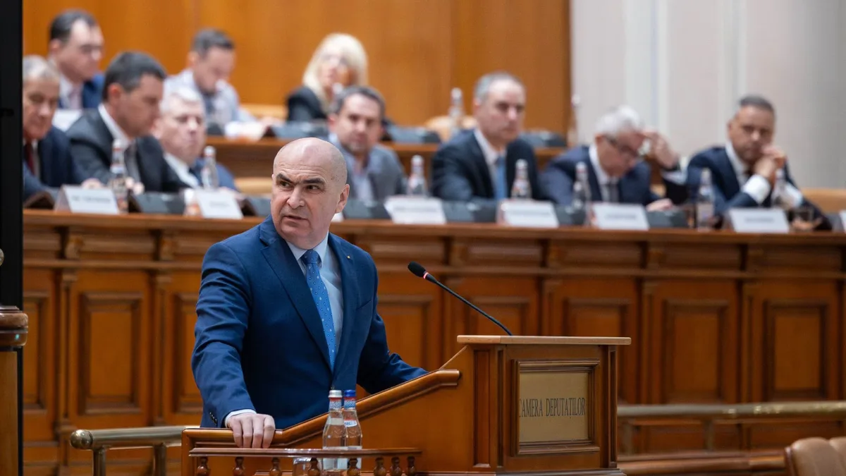 Ilie Bolojan la tribuna Camerei Deputaților, cu un microfon în față și participanți așezați în fundal.