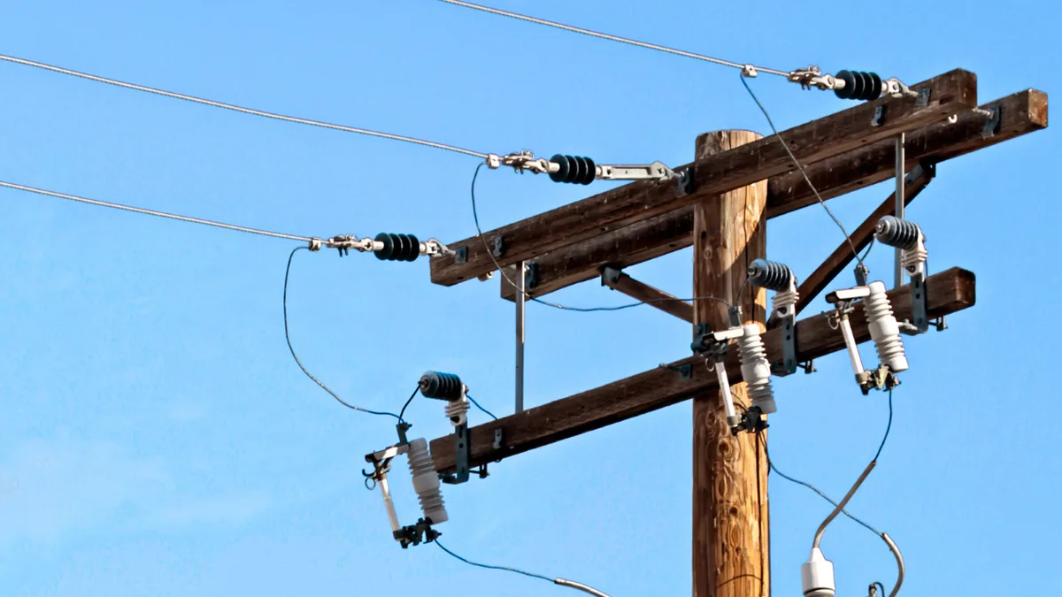 Stâlp din lemn cu traverse, conductoare electrice, izolatoare și echipamente de protecție, pe fundalul cerului albastru.