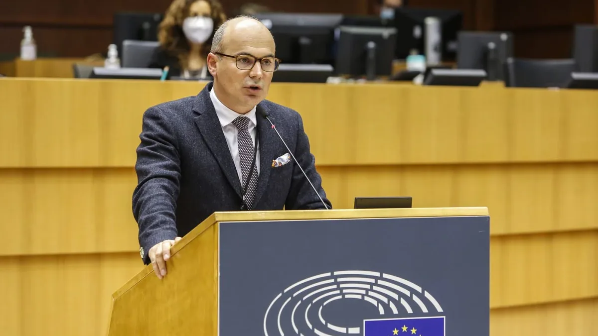 Rareș Bogdan vorbește la un pupitru cu emblema Parlamentului European, cu monitoare și participanți în fundal.