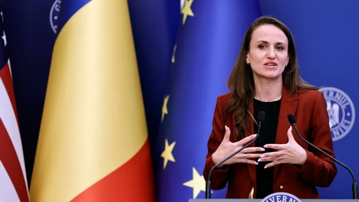 Oana Țoiu vorbește la două microfoane, în fața drapelelor României, Statelor Unite și Uniunii Europene.