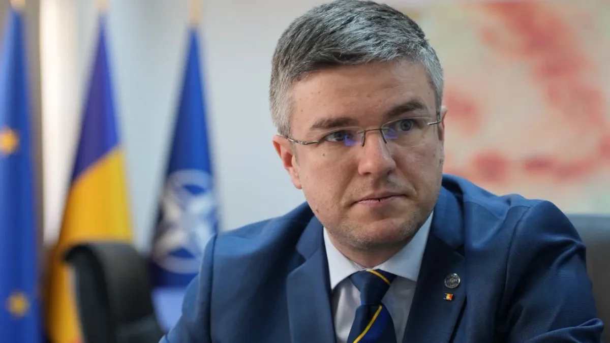 Irineu Darău, în costum albastru și cu ochelari, așezat în fața unor drapele.