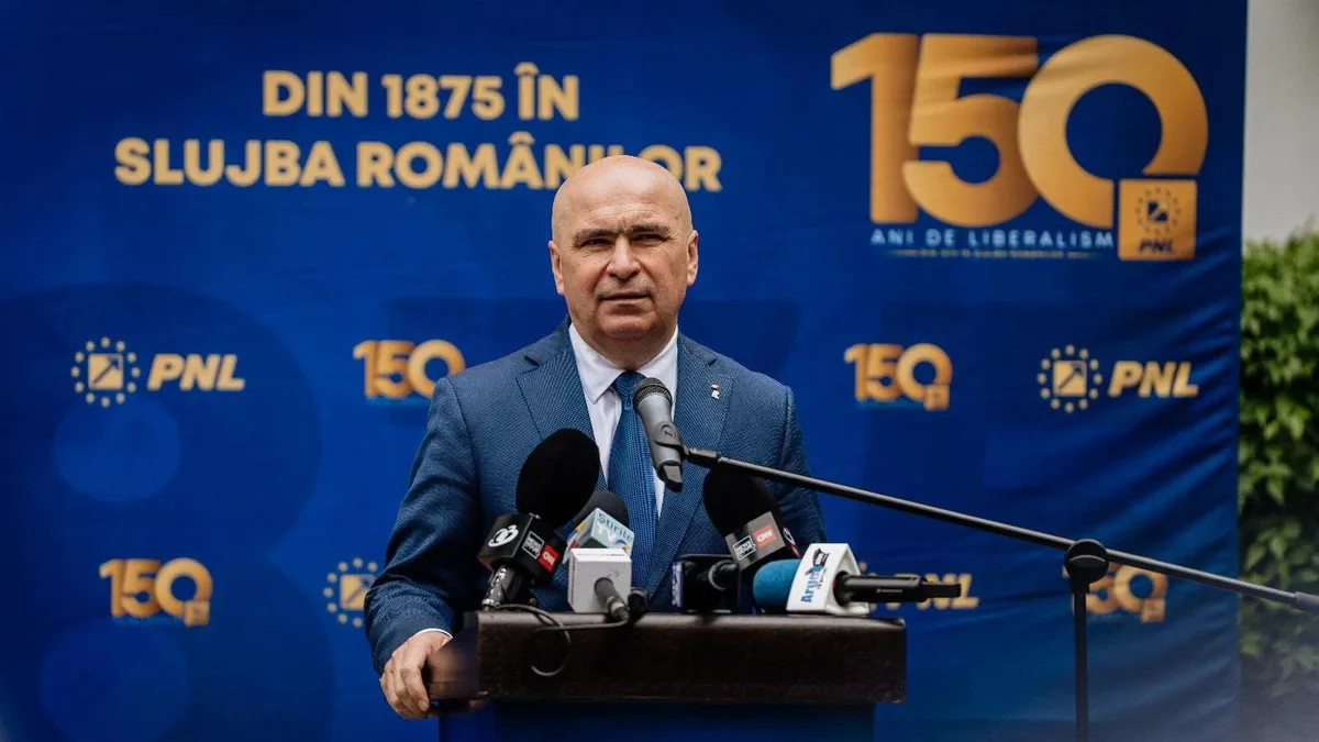 Ilie Bolojan la un pupitru cu microfoane, în fața unui panou albastru cu sigle PNL și însemne aniversare.